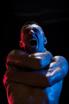 Unhappy shirtless sporty male shouting with eyes closed while embracing himself with arms crossed on chest and neck in light of red and blue lamps on black background