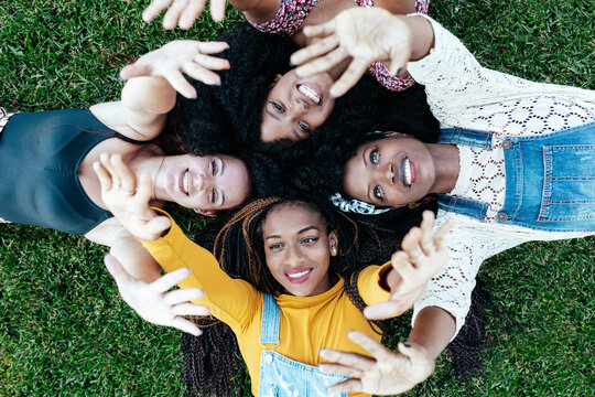 High angle of happy multiracial girlfriends lying together on grass and stacking hands while resting in park in summer