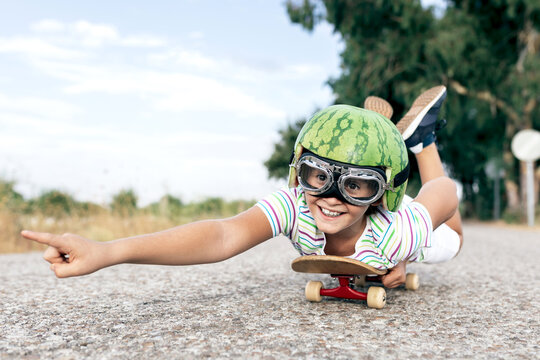 Ground level of happy kid in protective eyewear and ornamental watermelon helmet lying on skateboard on roadway and looking away