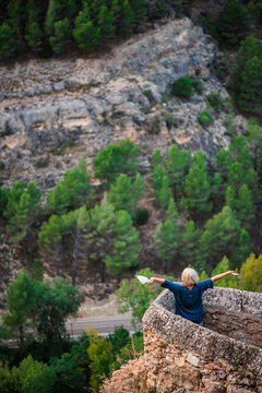 From above back view of female tourist with map standing near stone border of medieval fortress against rocky mountain slope with green forest and enjoying freedom while visiting Cuenca town in Spain