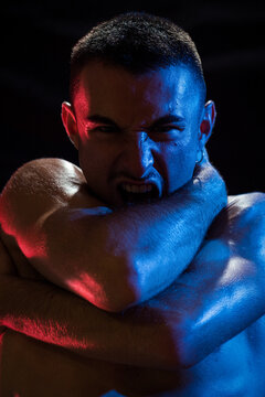 Unhappy shirtless sporty male shouting looking at camera while embracing himself with arms crossed on chest and neck in light of red and blue lamps on black background