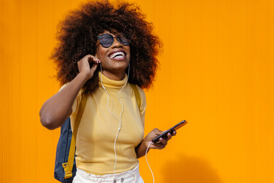 Black woman with afro hair listening to music on mobile in front of an orange wall