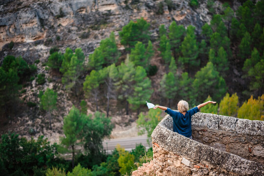 From above back view of female tourist with map standing near stone border of medieval fortress against rocky mountain slope with green forest and enjoying freedom while visiting Cuenca town in Spain
