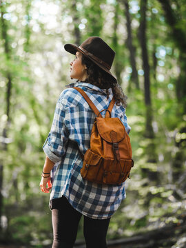 Back view of stylish traveling female with backpack and in checkered shirt standing in green woods and admiring nature during trip