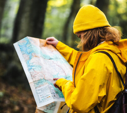 Side view of female explorer in yellow raincoat and with backpack standing in woods and searching for route on paper map