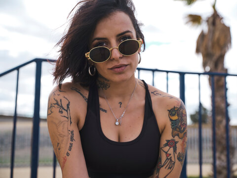Determined female millennial in street style wear and with tattoos sitting on ground in skate park and looking at camera while relaxing at weekend