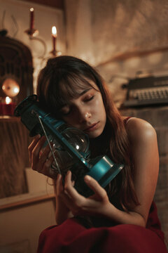 Sad female in red dress sitting in cozy room with old fashioned kerosene lantern during evening with eyes closed