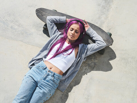 From above of confident female millennial with pink hair and in stylish clothes lying on longboard in skate park