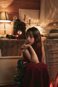 Peaceful female in red dress sitting in cozy room with old fashioned kerosene lantern while enjoying evening with eyes closed