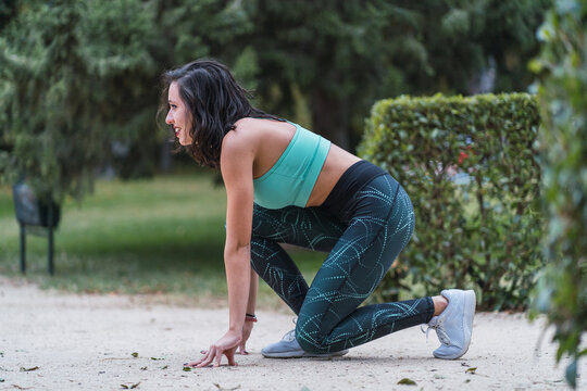 Side view of smiling female runner standing in crouch start position and getting ready for run during training in summer park