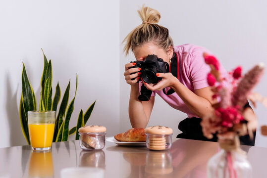Professional female photographer taking picture of desserts and glass of orange juice while working remotely from home