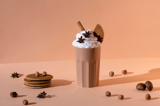 Glass with chocolate cocktail with whipped cream garnished with star anise and cinnamon stick placed on table with hazelnuts and cookies on pastel brown background
