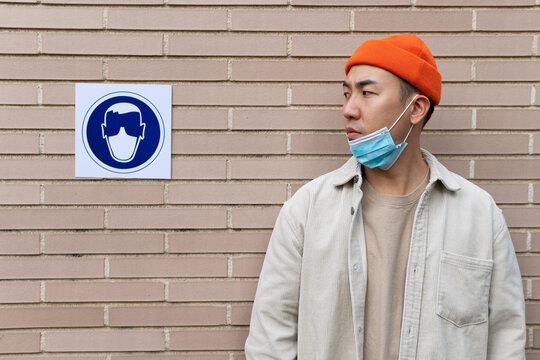 Serious Asian male wearing lowered mask looking at sign near building wall illustrating person in protective mask
