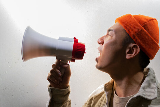 Side view of overwhelmed male protesting in casual clothes screaming in loudspeaker while standing against gray wall in dark room