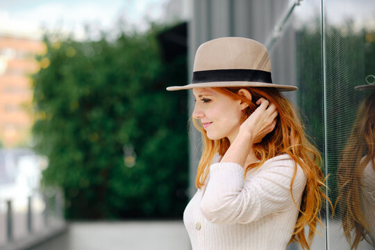 Side view of smiling young stylish ginger female in trendy hat touching hair while standing near modern building on city street