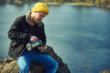Bearded adventurer man pouring tea or coffee from thermos in a metal cup while resting on a rock on automn nature background.