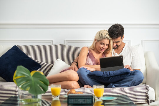 Happy young multiracial couple cuddling and browsing tablet while sitting together on cozy sofa near table with breakfast in morning - Powered by Adobe