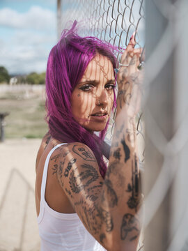 Informal female with long pink hair and tattooed arms leaning on metal grid in city and looking at camera