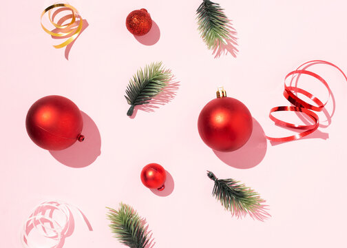 From above of red Christmas balls and green spruce branches arranged with shiny ribbons on pink background in studio