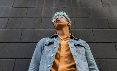 From below of young African American male model with blue hair and in casual outfit standing near urban building in city and looking away