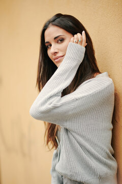 Charming female with long hair and in casual dress standing near building on street and modestly looking at camera