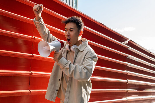 Agitating Asian male screaming in megaphone while standing with fist up near red metal fence and looking away in rage