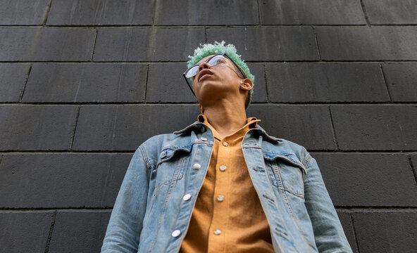 From Below Of Young African American Male Model With Blue Hair And In Casual Outfit Standing Near Urban Building In City And Looking Away