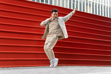 Full body joyful Asian male in casual wear listening to favorite song in wireless headphones while dancing along red metal fence