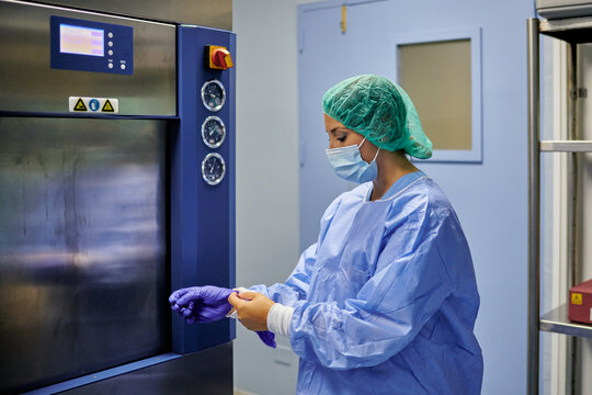 Side view competent female doctor wearing surgical uniform removing latex gloves while standing near sterilization machine in modern hospital