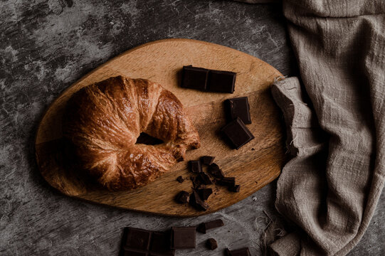 From above of delicious fresh crusty croissant and pieces of dark chocolate composed on wooden board on shabby table with linen fabric