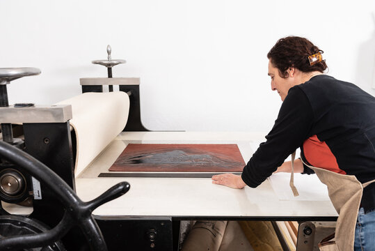 Focused middle aged female master putting sheet of linoleum with inked ornament on printing press while working in linocut technique in work studio