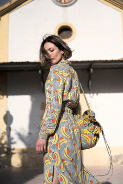 Pensive young gorgeous female with flying hair in dress with long sleeves looking down while standing next to old building with triangular roof in daytime