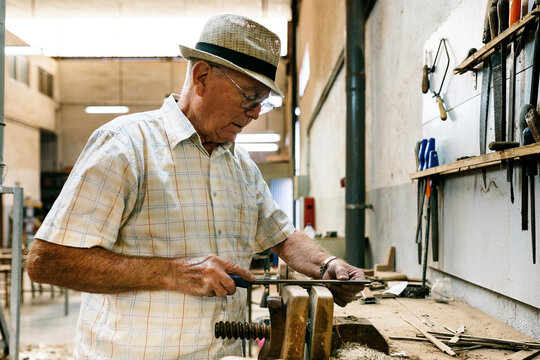 Side view of senior male carpenter grinding wooden details while making handmade folding fans in messy workshop