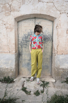 Full body of faceless female in shirt with floral print and light green trousers looking down while standing at shabby door of old construction in daytime