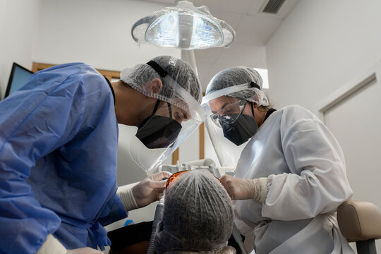 Professional stomatologist with assistant wearing protective masks and face shields sitting near patient with expander in mouth in modern clinic