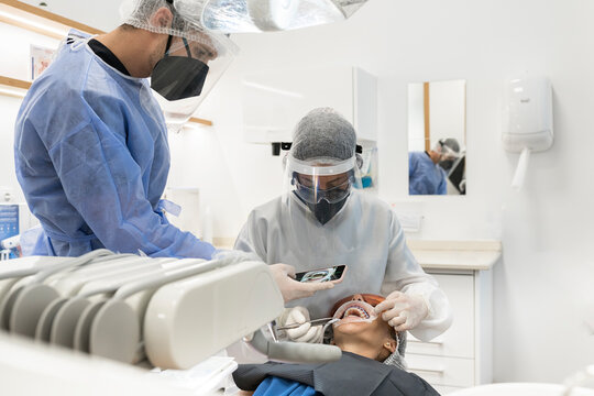 Male dentist in protective mask and face shield taking picture of teeth of patient on smartphone after performing healing procedure while working with colleague in modern dental room