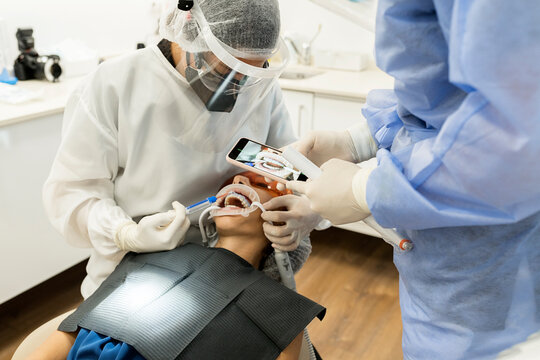 Male dentist in protective mask and face shield taking picture of teeth of patient on smartphone after performing healing procedure while working with colleague in modern dental room