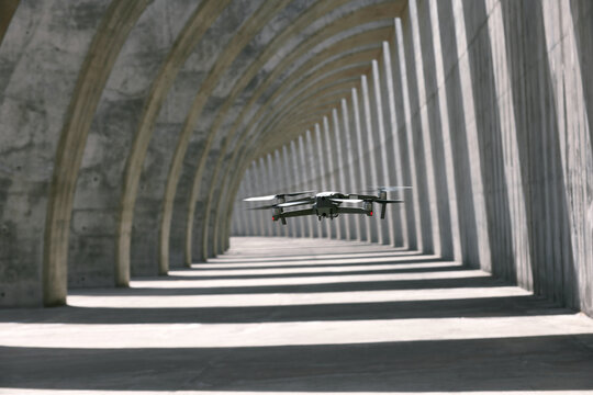 Modern drone flying in empty arched walkway with columns casting shadows in shape of lines on sunny day