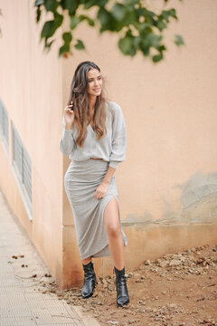 Charming female with long hair and in casual dress standing near building on street and modestly looking away