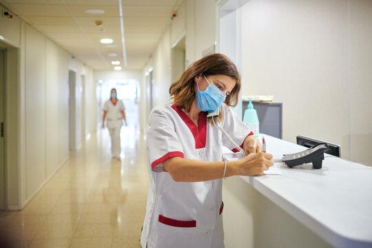 Concentrated female nurse wearing white medical suit and face mask taking notes in clinical record chart while standing near modern hospital reception