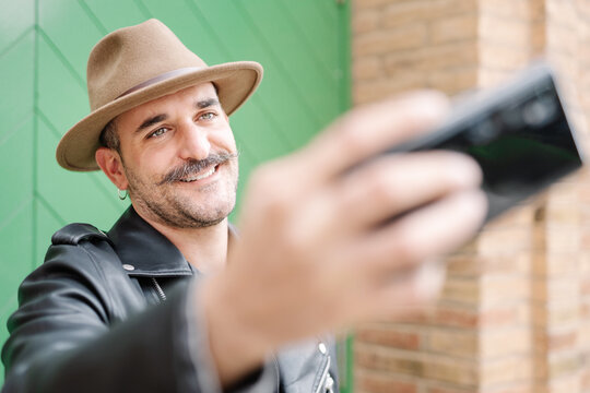 Cheerful glad bearded gallant male in hat taking photo in yard of brick house
