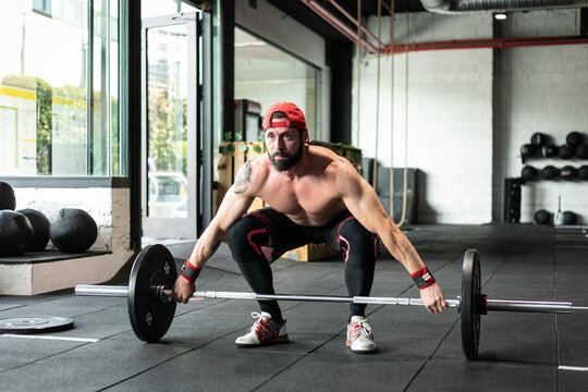 Muscular focused shirtless male athlete doing clean and jerk exercise with barbell during weightlifting training in gym looking away