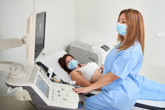 Side view of female doctor in uniform examining patient in mask while watching ultrasound scanner monitor in clinic