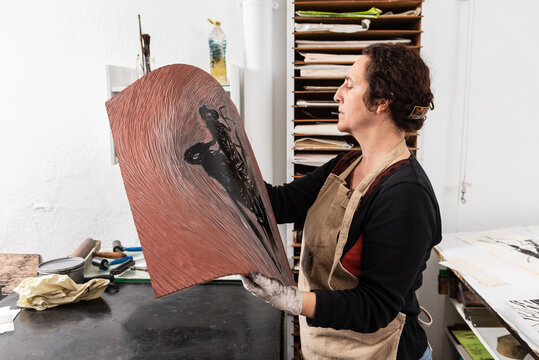 Side view of concentrated middle aged craftswoman holding piece of linoleum with inked pattern while making print in creative workplace