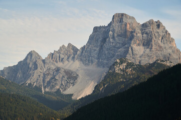 Picturesque scenery of high mountain in Dolomites under clouds near green coniferous forest under cloudy bright sky