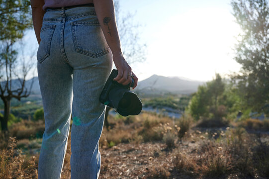 Back view of crop unrecognizable female hiker with professional photo camera standing on grassy hill and admiring picturesque scenery of autumn forest and mountains