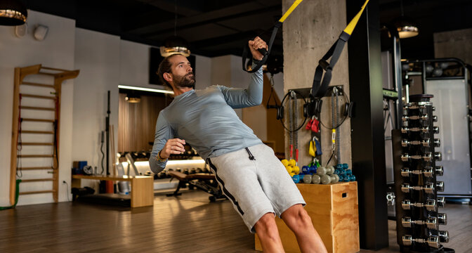 Side view of concentrated muscular male athlete practicing suspension training with TRX ropes in modern gym