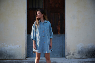Young slim female in stylish denim dress and boots standing on grassy lawn covered with hoarfrost against weathered rural house in autumn day