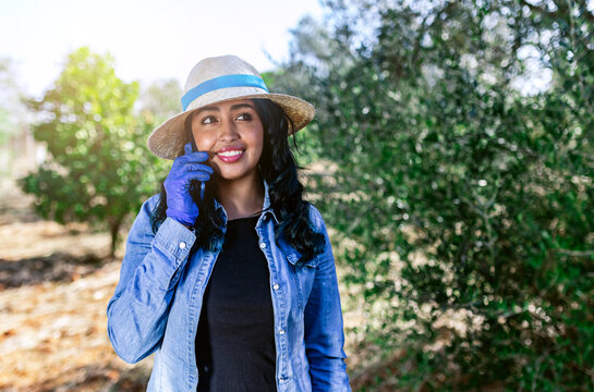 Smiling young female talking on phone while working in green garden with fruit trees
