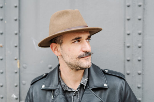 Trendy thoughtful male in hat and leather jacket looking away standing on metal door background